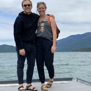 two people in black standing on a dock in front of some gorgeous aqua water with a little hill on the horizon