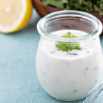 a little jar of ranch dressing. white and creamy with an unnatural pile of parsley on top. Half a lemon in the background next to a wooden bowl of curly kale