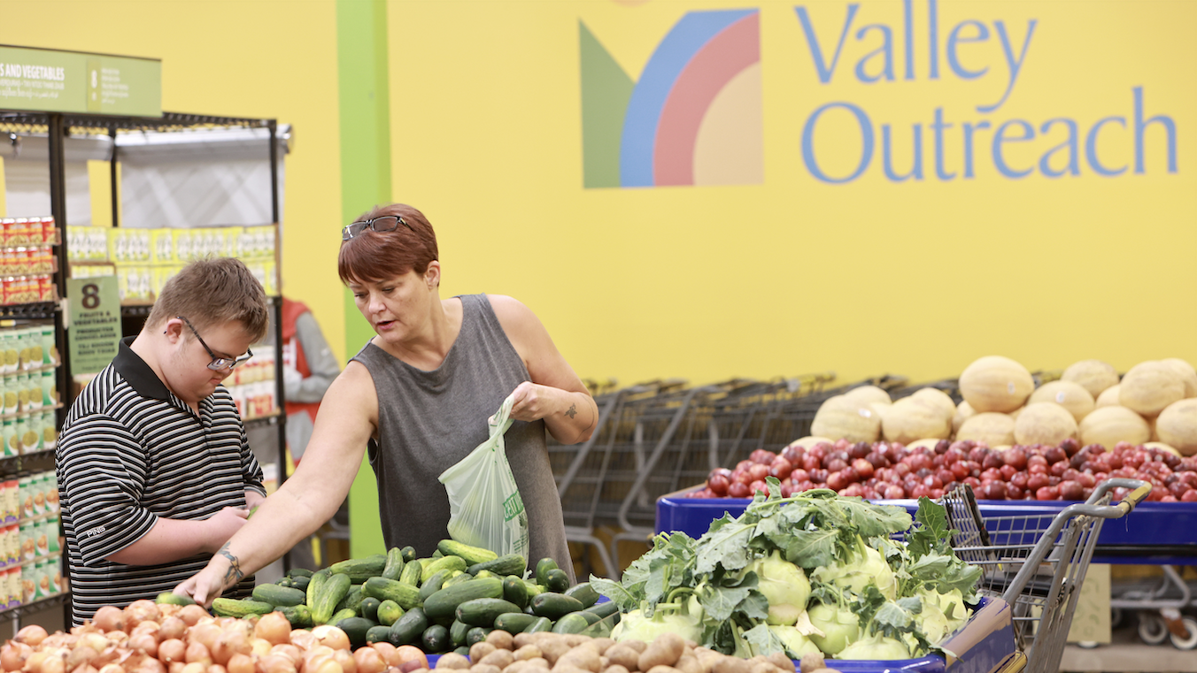 two people picking out fresh produce at valley outreach food shelf. walls are yellow, the produce they're choosing from are bright green cucumbers, yellow onions, and. . . some large green kohlrabi! A table of red potatoes and cantaloupe melons is in the background 
