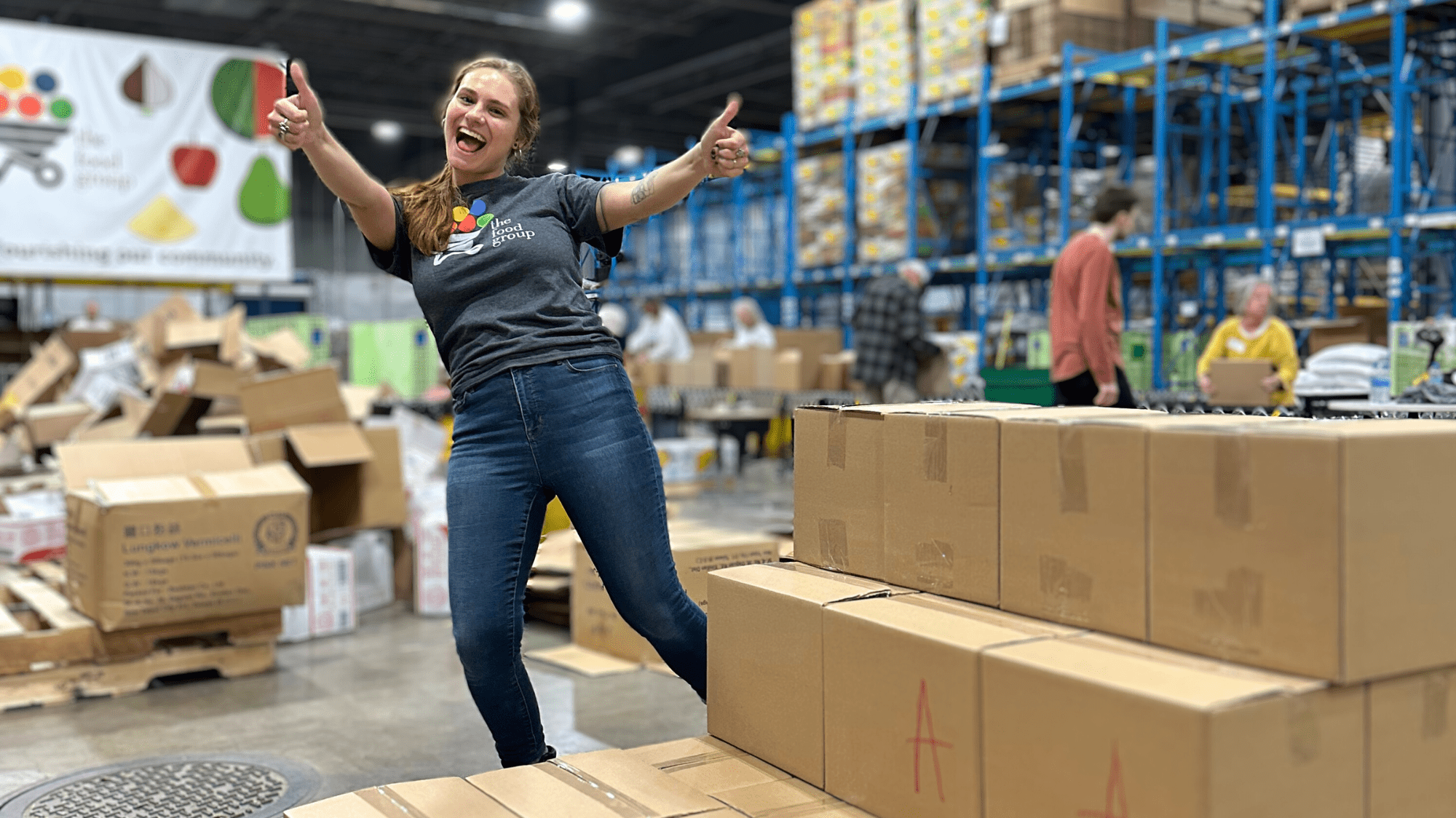 volunteer leader smiling and giving two thumbs up. they're in a warehouse next to a huge stack of cardboard boxes packed with food!