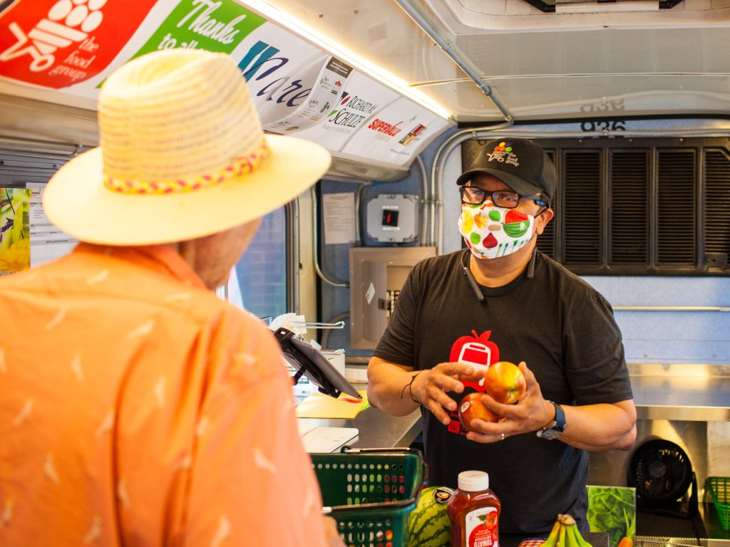 An exchange between two gentlemen on a bus at the Twin Cities Mobile Market. The man on the left is wearing a fedora and a bright orange shirt with his back facing the camera. The other man is wearing a black cap, a mask, and a black t-shirt, and he is holding two apples looking at the man in orange. A shopping basket, watermelon, ketchup, and bananas are on the counter.