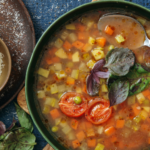 bowl of tomato soup with veggie chunks in it. blue background and spoon in bowl