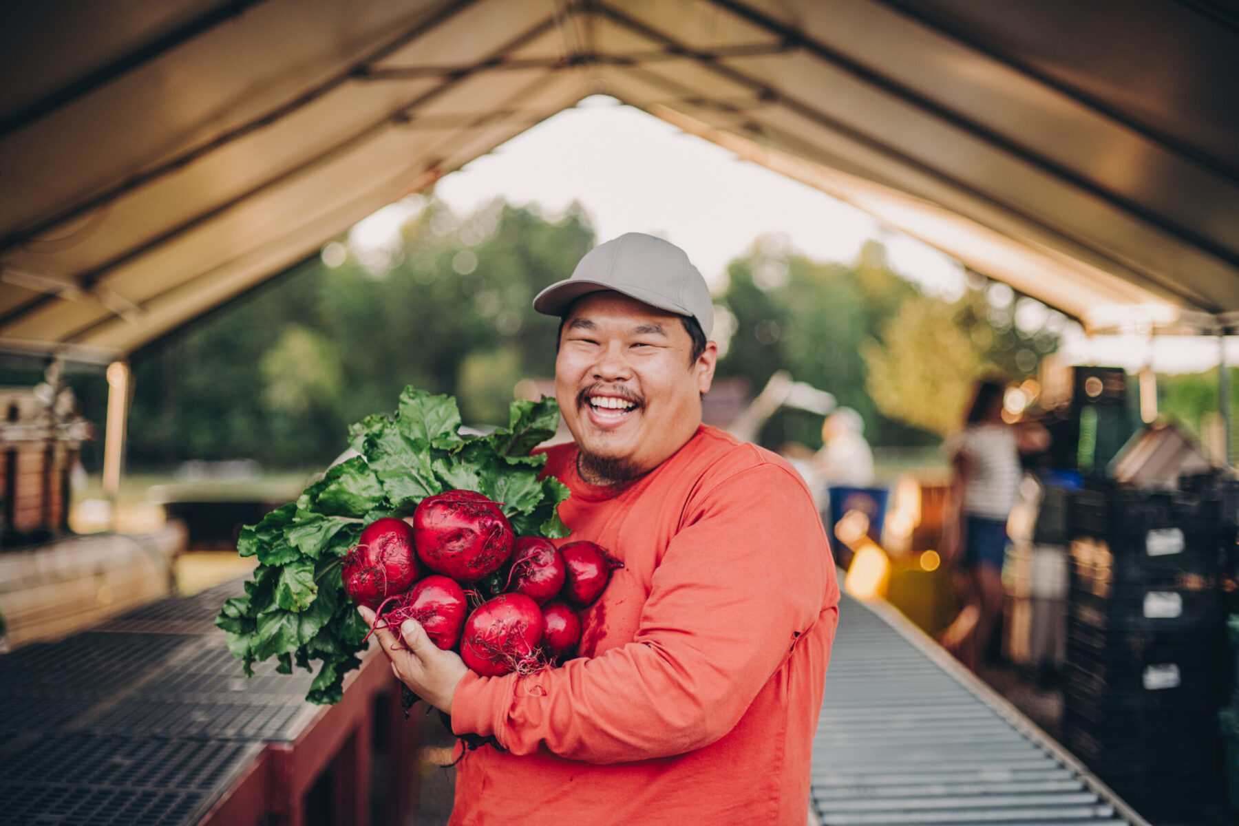 A cheerful looking male Asian American farmer smiles as he holds a bunch of bright red beets.