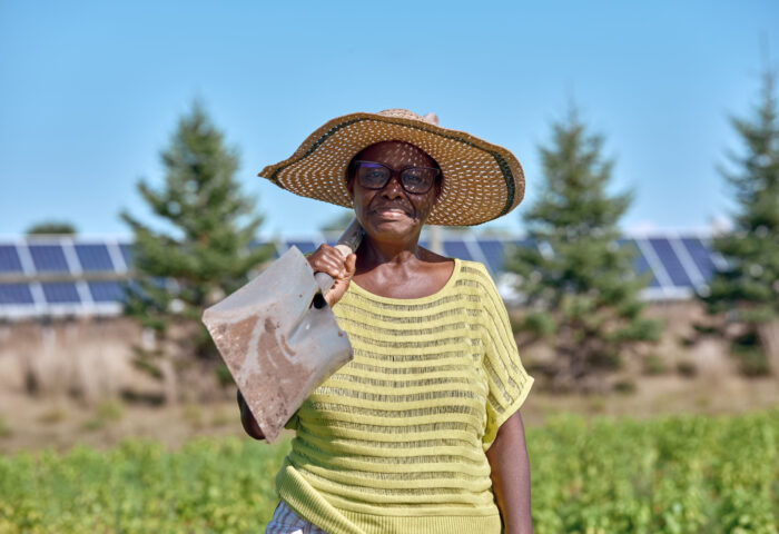 A woman with a shovel stands smiling with a shovel in her field at our agrisolar farm