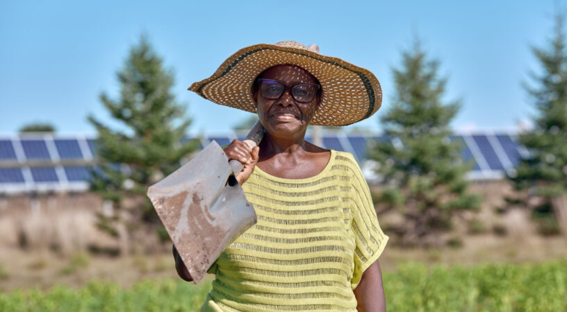 A woman with a shovel stands smiling with a shovel in her field at our agrisolar farm