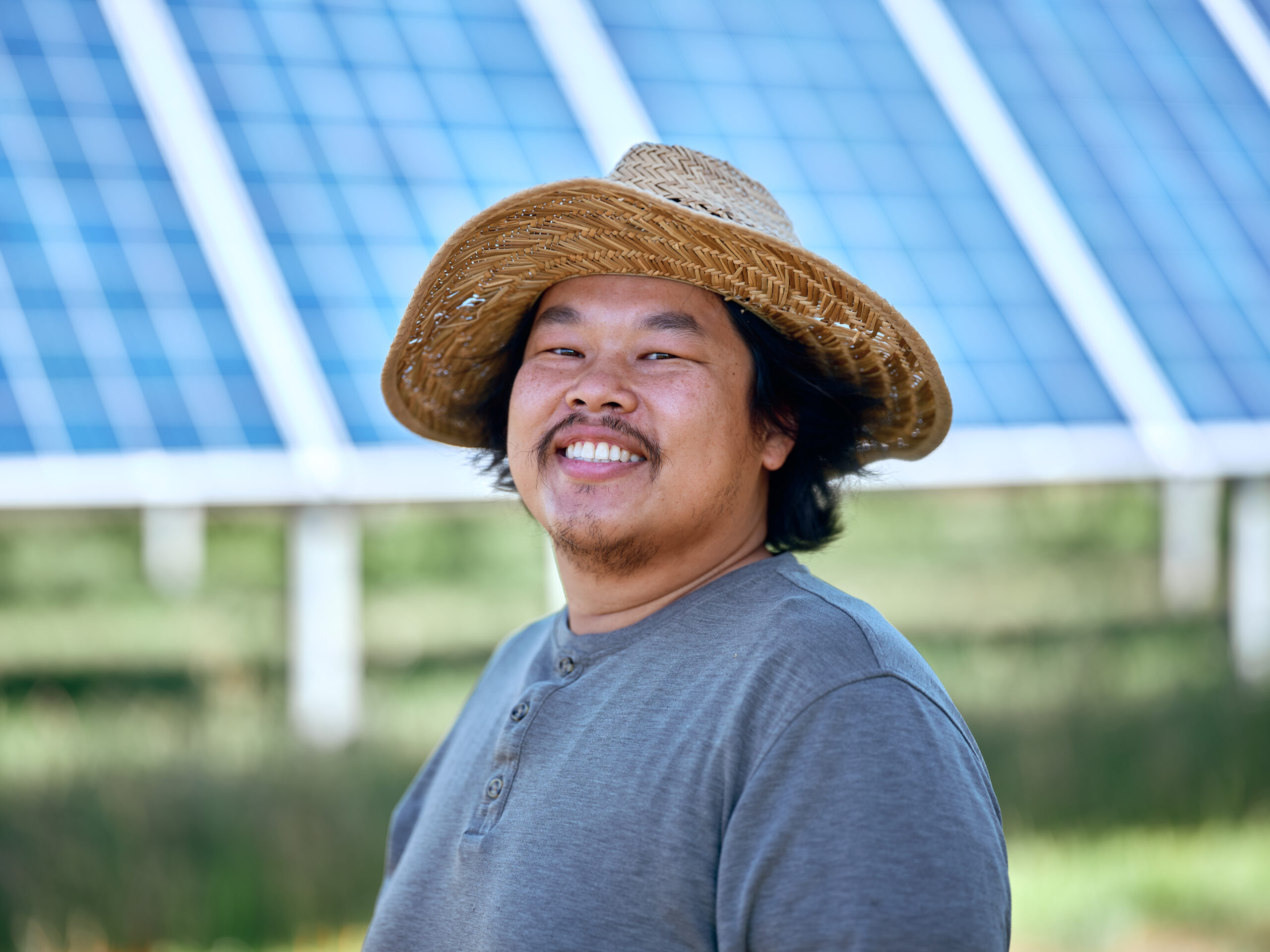 A farmer in a straw hat stands and smiles in front of a solar panel
